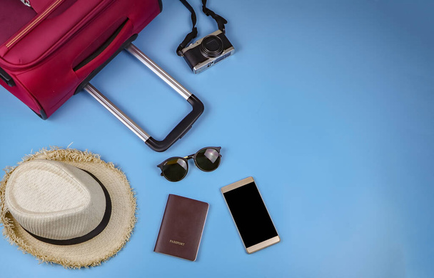 Conjunto plano de accesorios de viajero, pasaporte, sombrero, teléfono móvil, cámara, gafas de sol y maleta rosa sobre fondo azul. concepto de viaje
. - Foto, Imagen