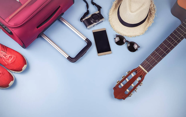 Płaskie ułożenie akcesoriów podróżniczych, kapelusz, telefon komórkowy, gitara, aparat fotograficzny, okulary przeciwsłoneczne, trampki i różowa walizka na niebieskim tle. koncepcja podróży. - Zdjęcie, obraz