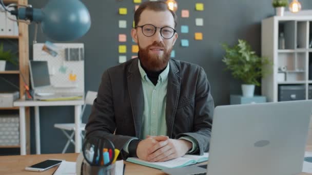 Portret młodego przedsiębiorcy przy biurku w biurze patrzącego w kamerę z lekkim uśmiechem - Materiał filmowy, wideo
