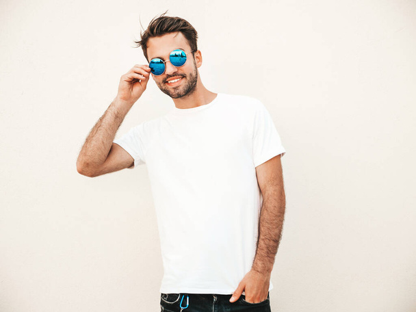 Portrait of handsome smiling stylish hipster lambersexual model.Man dressed in white T-shirt. Fashion male posing on the street background near wall in round sunglasses - Foto, afbeelding