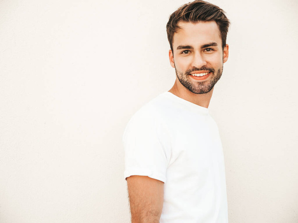 Retrato de guapo sonriente modelo hipster lambersexual.Hombre vestido con camiseta blanca. Moda masculina posando en el fondo de la calle cerca de la pared en gafas de sol redondas - Foto, Imagen