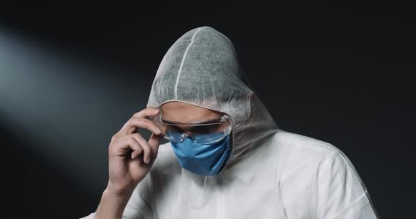 Portré fáradt kaukázusi férfi fiatal orvos teljesen felszerelt jelmez és orvosi maszk figyelembe szemüveg és nézi a kamera sötét háttérrel. A covid-19 koncepció globális járványa Laboratóriumi dolgozó - Felvétel, videó