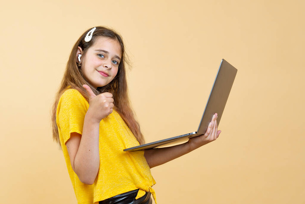 Портрет улыбающейся юной брюнетки в беспроводных наушниках, держащей ноутбук и показывающей хорошо жест изолирован на желтом фоне
 - Фото, изображение