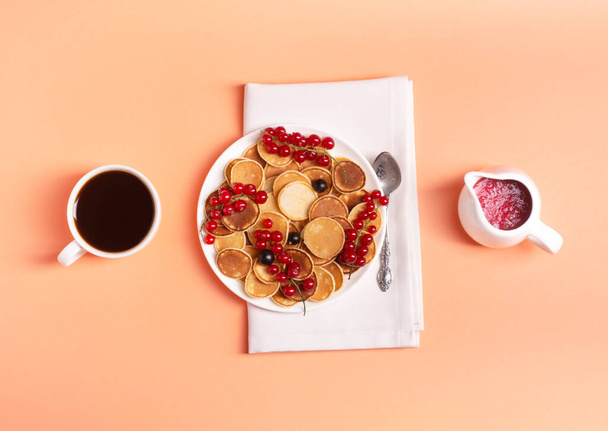 Hausgemachte Mini-Beutel in einem weißen Teller mit roten Johannisbeeren auf Pfirsichhintergrund, neben einer Tasse Espresso und roter Johannisbeermarmelade, flache Lage, ein leckeres Frühstückskonzept. - Foto, Bild