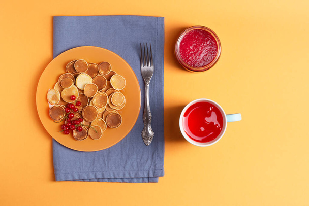 Tasty mini pancakes in an orange plate stand on a blue napkin, next to it is a blue mug with berry drizzle and a jar of jam, orange background, knolling, flat lay, horizontal orientation. - Photo, image