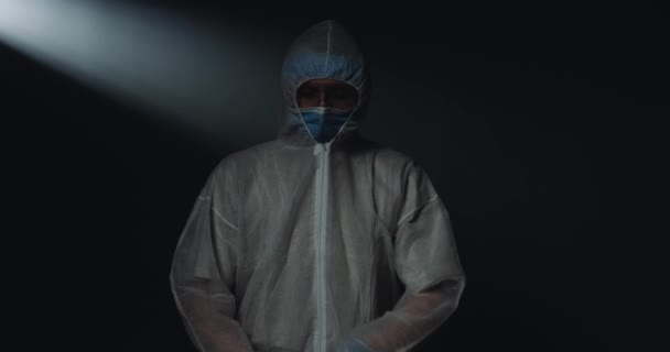 Portrait of Caucasian male young doctor epidemiologist in full protection costume with medical mask and goggles looking at camera with tired eyes on black background. Medic during coronavirus epidemic - Materiał filmowy, wideo