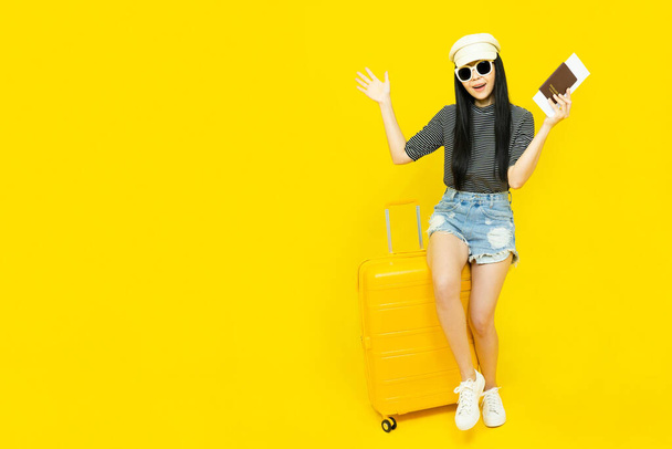 Young Asian female tourist in casual clothes and sunglasses Carrying a  passport to go to travel sitting on a luggage isolated on yellow background copy space,Vacation,holidays and travel concept - Fotografie, Obrázek