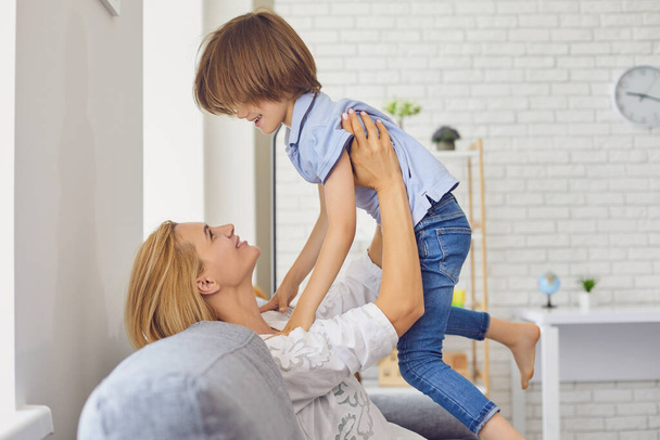 Happy mother lifting her son up into air at home. Parent playing game with little child, having fun in living room - Φωτογραφία, εικόνα