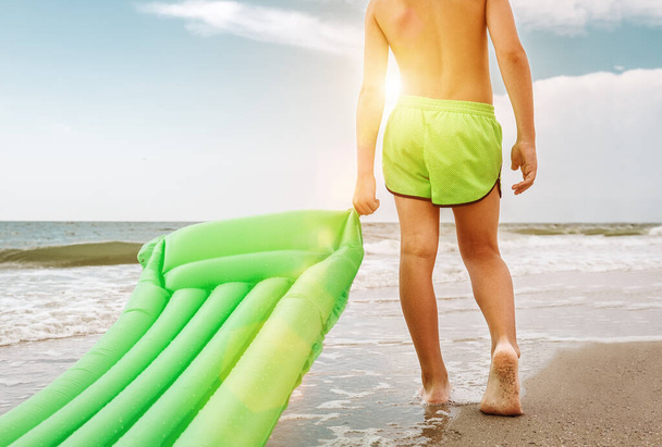 Boy with green swimming mattress stands on seasurf line - 写真・画像