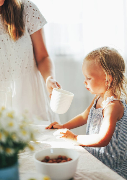 かわいいブロンドの幼児の女の子は、晴れた日にテーブルで母親の手からカップから飲む。コテージコア美学 - 写真・画像