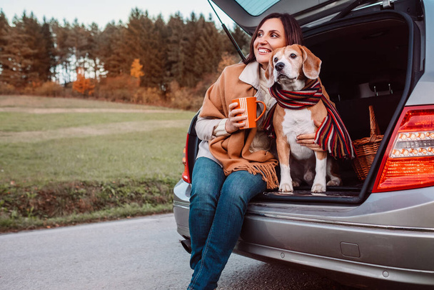 Woman and dog with shawls sits together in car trunk  on autumn road - Zdjęcie, obraz