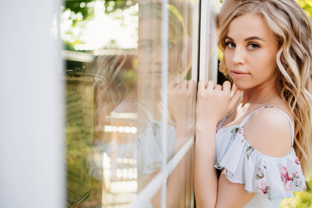 beautiful blonde girl in summer dress standing by  window in garden in backyard. - Photo, image