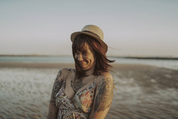 A young female with tattoos wearing a dress and straw hat on blurred ocean background - Fotografie, Obrázek