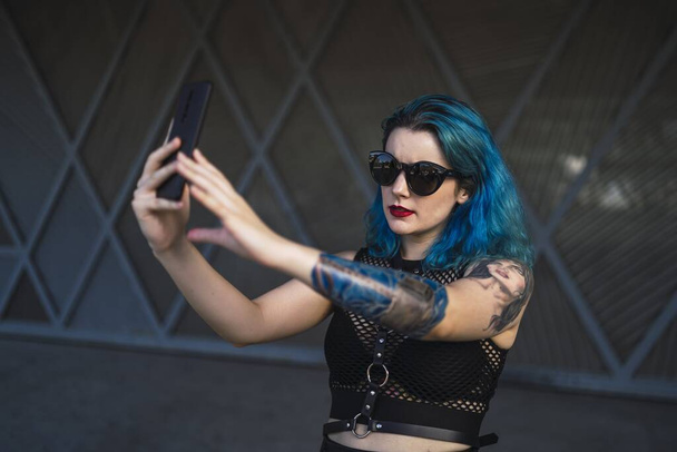 A europian female with tattoo and blue hair wearing sunglasses and sexy black dress taking a selfie at the side of a building - Foto, Imagem