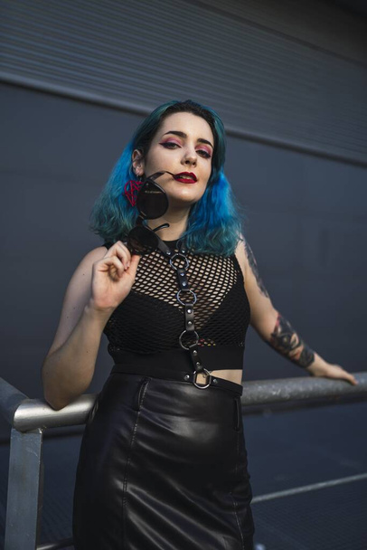A European female with tattoo and blue hair wearing sunglasses and sexy black dress posing on metal railings at the side of a building - Фото, зображення