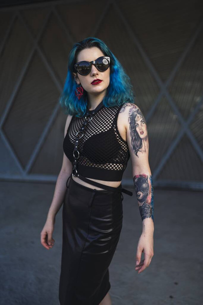 A European female with tattoo and blue hair wearing sunglasses and sexy black dress posing at the side of a building - Zdjęcie, obraz
