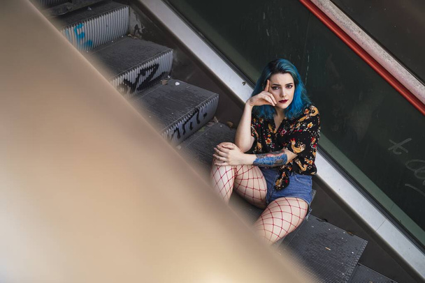 A young female with blue hair wearing shorts sitting on an escalator under the sunlight - Fotografie, Obrázek