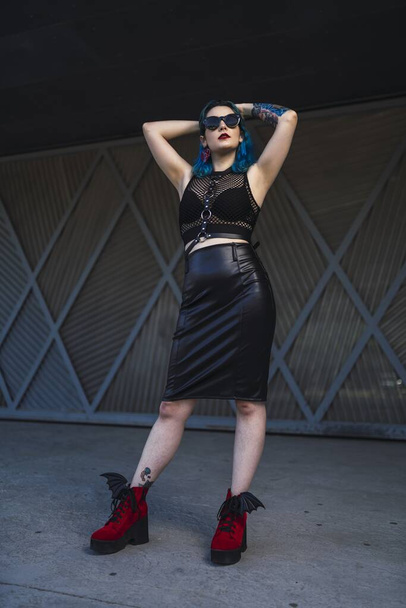 A European female with tattoo and blue hair wearing sunglasses and sexy black dress posing at the side of a building - Фото, изображение