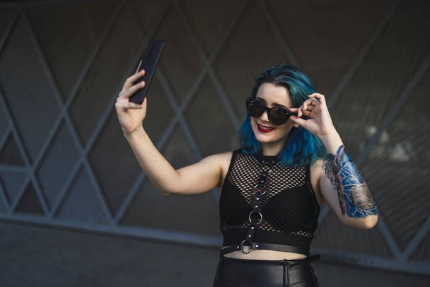Una hembra europea con tatuaje y pelo azul usando gafas de sol y vestido negro sexy tomando una selfie al lado de un edificio - Foto, imagen