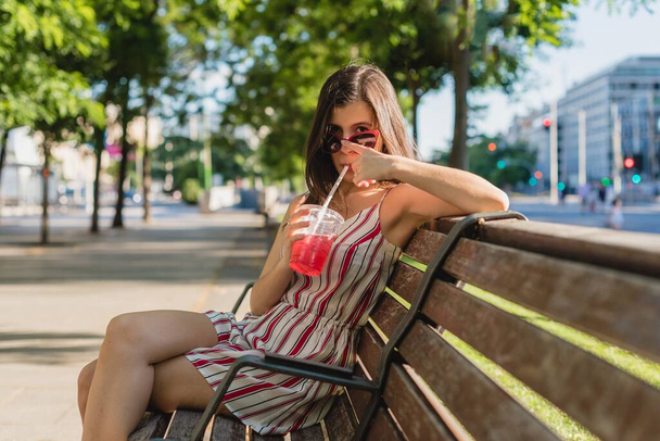 A young girl with stripped dress and sunglasses drinking juice on the bench - 写真・画像