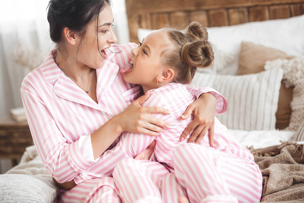 Young active mother and her little daughter having fun together at home in the bedroom wearing pajamas - Foto, Bild
