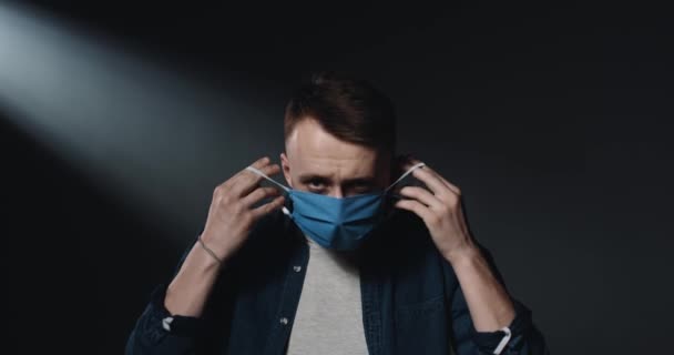 Portrait shot of Caucasian young handsome guy taking on medical mask and looking at camera with sad face. Dark wall background. Upset male patient. Coronavirus pandemic concept. - Filmagem, Vídeo
