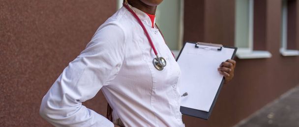 African American Nurse black young woman doctor, in a white coat, with a phonendoscope, holds a folder for medical papers in a rue, against the background of a hospital. A young black girl of twenty seven years old, a doctor in a white coat, smiles,  - Фото, изображение
