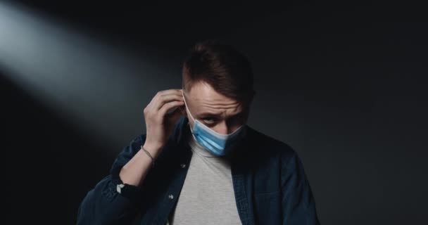 Portrait of Caucasian young handsome guy taking on blue medical mask and looking at camera with serious sad face on black wall background. Coronavirus pandemic. Safety concept. - Felvétel, videó