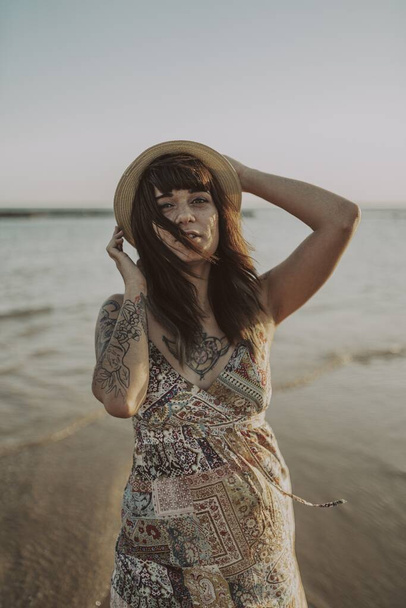Bulanık okyanus arka planında elbise ve hasır şapka takan genç bir kadın. - Fotoğraf, Görsel