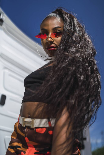 A shallow focus of an African female with long hair wearing a crop top and red camouflage trousers under the sunlight - Foto, imagen