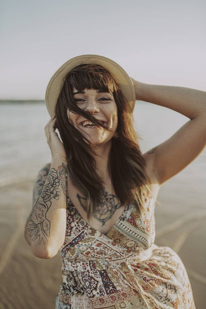 A young female with tattoos wearing a dress and straw hat on blurred ocean background - Фото, зображення