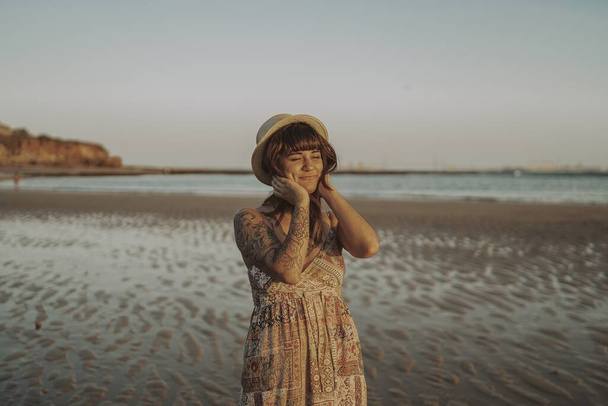 A young female with tattoos wearing a dress and straw hat on blurred ocean background - Zdjęcie, obraz