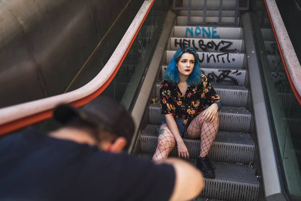 A Caucasian female with blue hair posing on an escalator and a photographer taking her picture - Фото, зображення