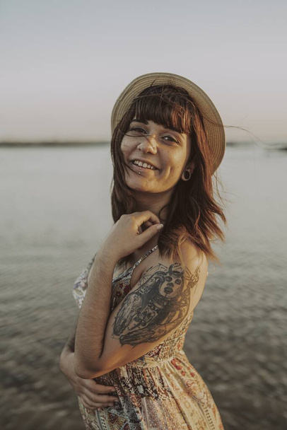 A young female with tattoos wearing a dress and straw hat on blurred ocean background - Foto, imagen