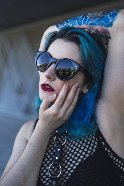 A European female with tattoo and blue hair wearing sunglasses and sexy black dress posing at the side of a building - Фото, зображення