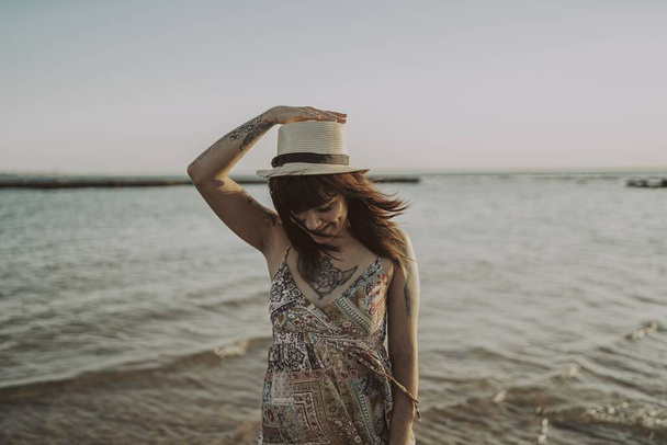 A young female with tattoos wearing a dress and straw hat on blurred ocean background - Fotó, kép