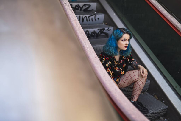 A young female with blue hair wearing shorts sitting on an escalator under the sunlight - Zdjęcie, obraz
