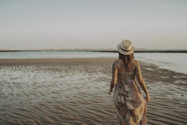 Una joven hembra con tatuajes con vestido y sombrero de paja sobre fondo borroso del océano - Foto, Imagen