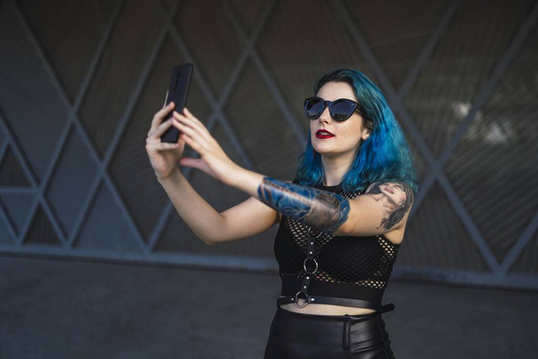 A europian female with tattoo and blue hair wearing sunglasses and sexy black dress taking a selfie at the side of a building - Фото, изображение