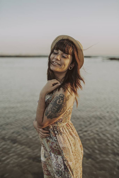 A young female with tattoos wearing a dress and straw hat on blurred ocean background - Foto, Bild