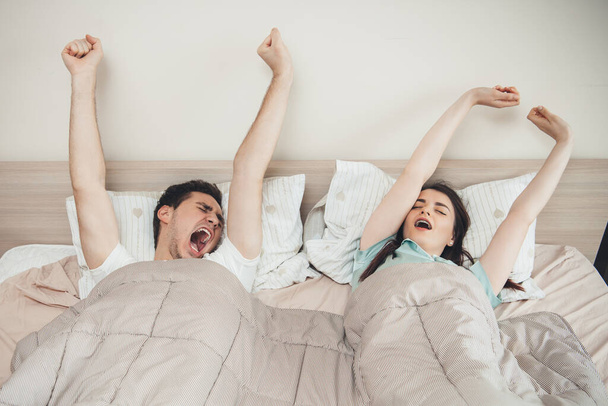 ベッドの中で午前中に白人のカップルがストレッチと掛け布団で覆われて立ち上がる - 写真・画像