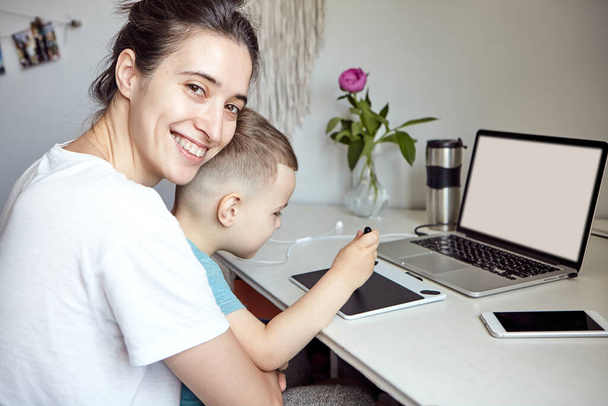 Egy nő egy gyerekkel egy számítógépnél dolgozik. Fogalom a munka az otthoni és otthoni családi oktatás. Anya és fia egy grafikus tableten és egy laptopon dolgoznak otthon.. - Fotó, kép