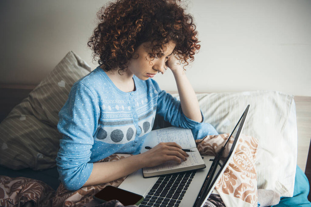 Λευκή γυναίκα με σγουρά μαλλιά διαβάζει το σημειωματάριό της πριν από τις online εξετάσεις στο κρεβάτι με ένα φορητό υπολογιστή - Φωτογραφία, εικόνα