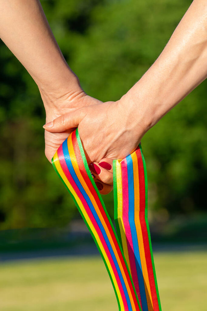 LGBT pride concept. Women hold each other hand and LGBT ribbon in front of the green nature background. Hands is in camera focus - Photo, Image