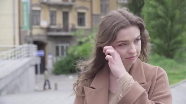 Close-up of charming brunette Caucasian woman crying outdoors on windy day. Portrait of beautiful young lonely girl standing on city street alone. Depression, sadness, negative emotions. - Video, Çekim