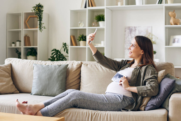 超音波画像を持つ幸せな妊娠中の女性彼女の赤ちゃんは、リビングルームでソファに座っている間、スマートフォンのカメラの前で自画自身を作ります - 写真・画像