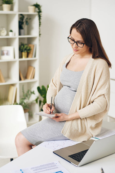 Досить елегантний вагітний працівник білого комірця сидить на столі за ноутбуком і записує план на день в блокноті в офісі
 - Фото, зображення