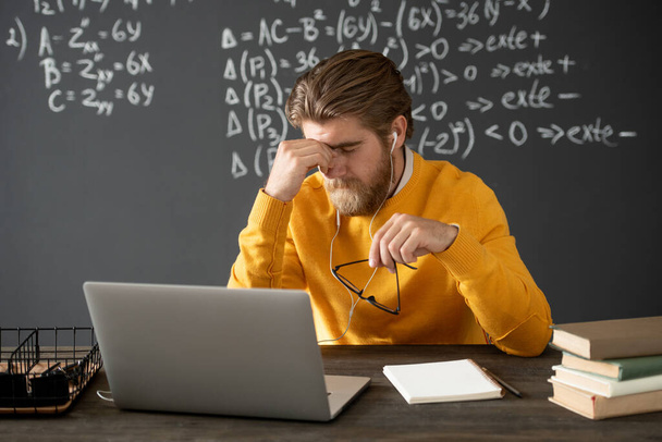Tired algebra teacher in casualwear touching his face while bending over table in front of laptop during online lesson against blackboard - Photo, Image