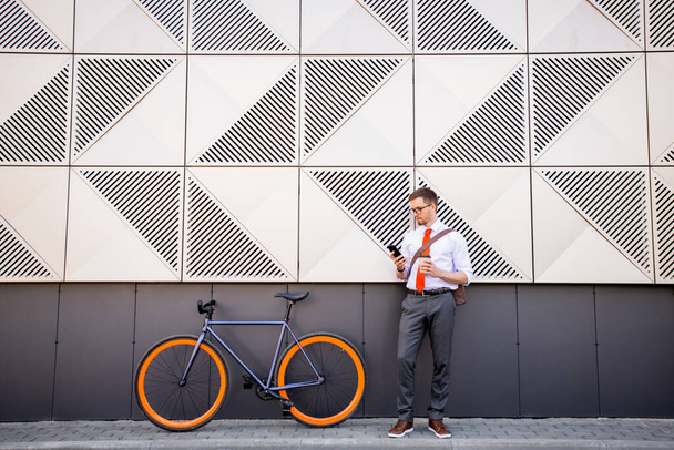 Спокійний молодий бізнесмен, використовуючи смартфон і каву на перерві, стоячи на стіні сучасного бізнес-центру
 - Фото, зображення