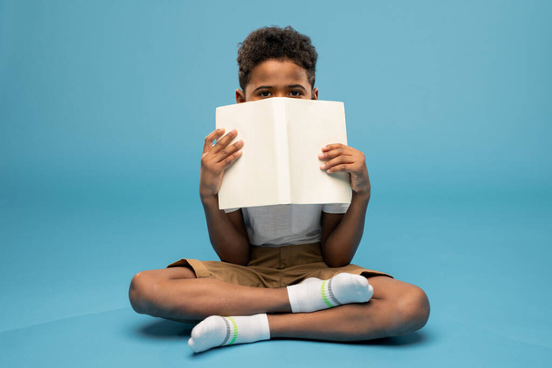 Schattig klein schooljongetje van Afrikaanse etniciteit houdt het boek dicht bij zijn gezicht en gluurt uit zijn cover terwijl hij op de vloer zit - Foto, afbeelding